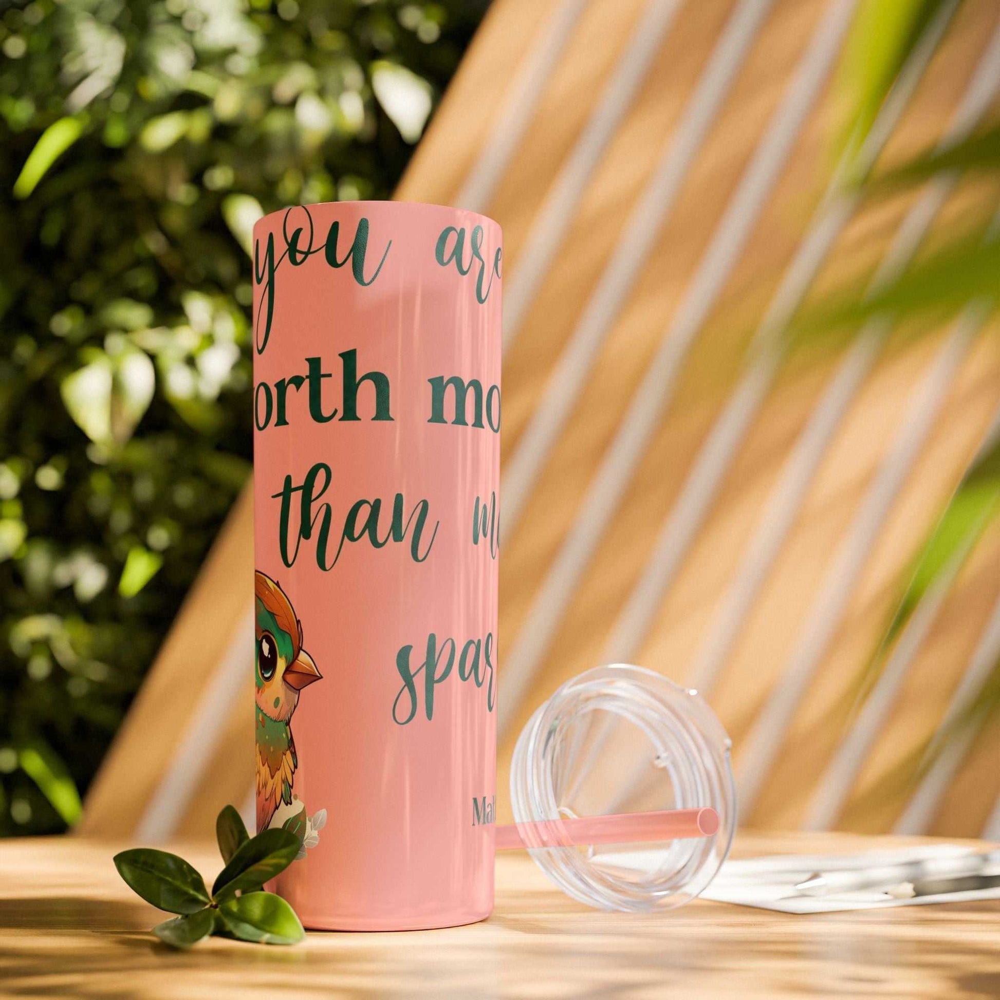 Stainless steel tumbler with "Worth More Than Sparrows" design, 20oz, pink with lid and straw, outdoors.