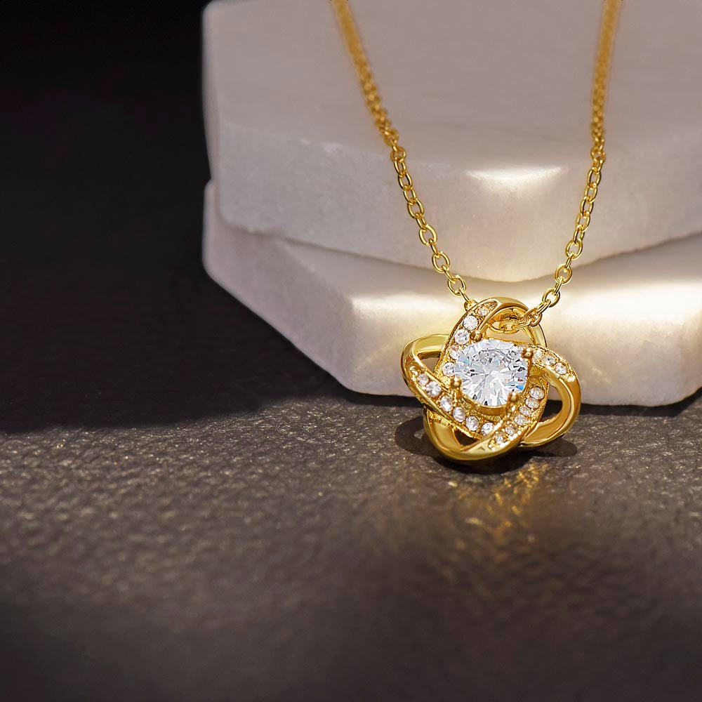 Love Knot necklace with cubic zirconia pendant, 18k gold over stainless steel, displayed on a textured surface.