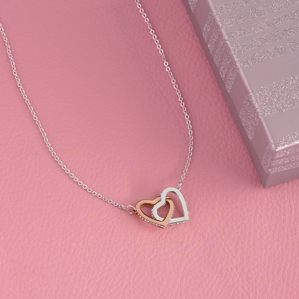 Interlocking hearts necklace with cubic zirconia on a pink background, near a gift box.